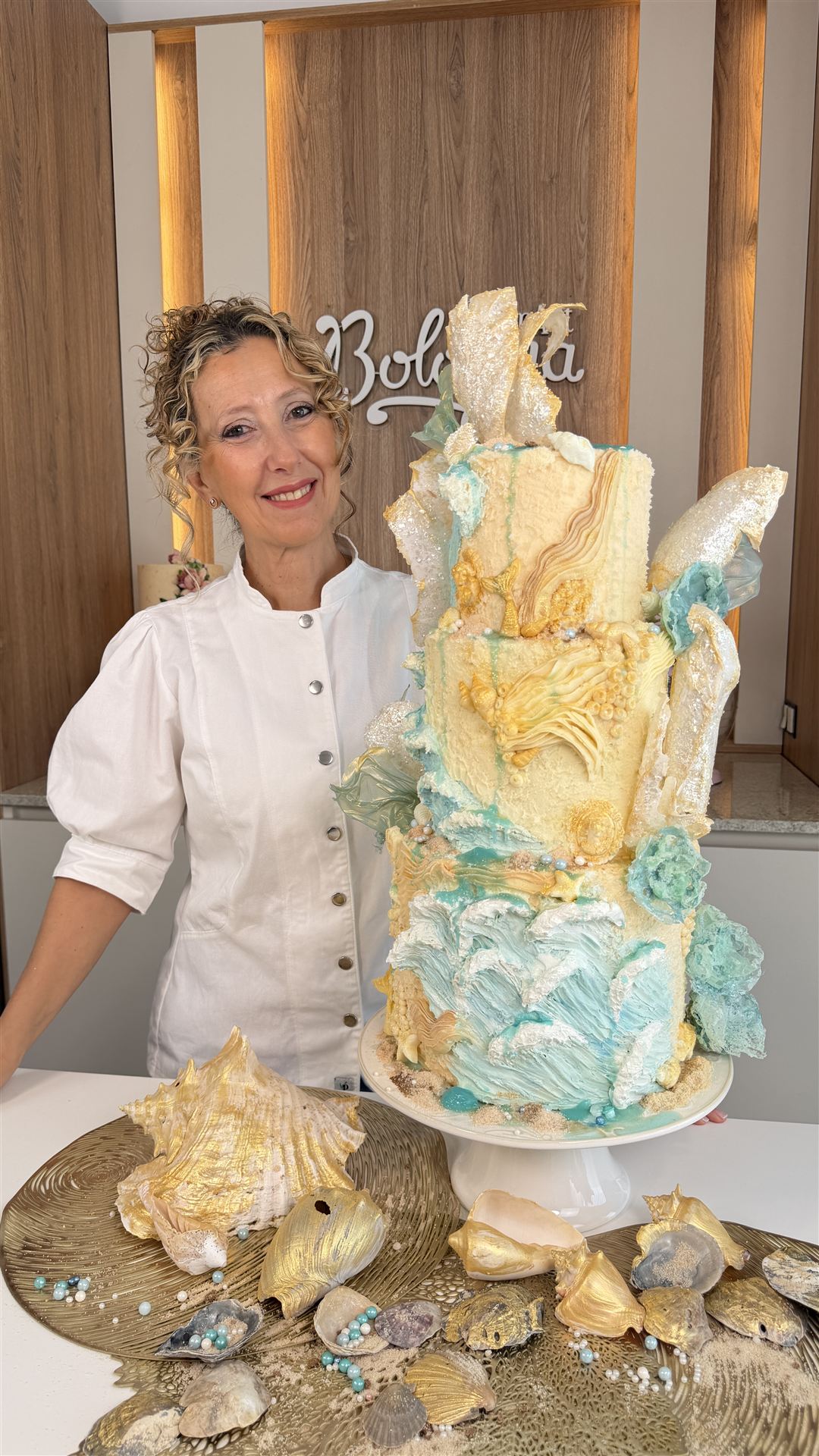 Adriana Bologna sonriendo junto a su pastel de tres pisos con olas de buttercream turquesa, caracolas doradas y velas de azúcar